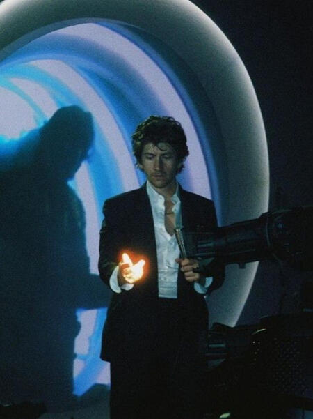alex turner stands on a stage tinted with blue light, holding his hand, which glows lit by a spotlight, in front of him
